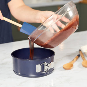 Someone pours cake batter from a glass bowl into the Zyliss Durable Non-Stick Springform Cake Pan using a blue spatula on a light countertop, with wooden spoons nearby. #20cm/8"