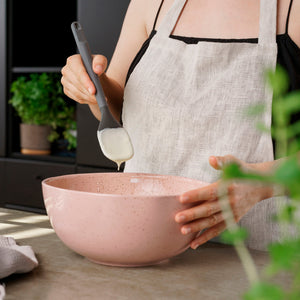 Wearing a light apron, a person uses a Zyliss Spoontula to mix creamy white batter in a pink bowl, with green plants visible in the kitchen background. #Medium