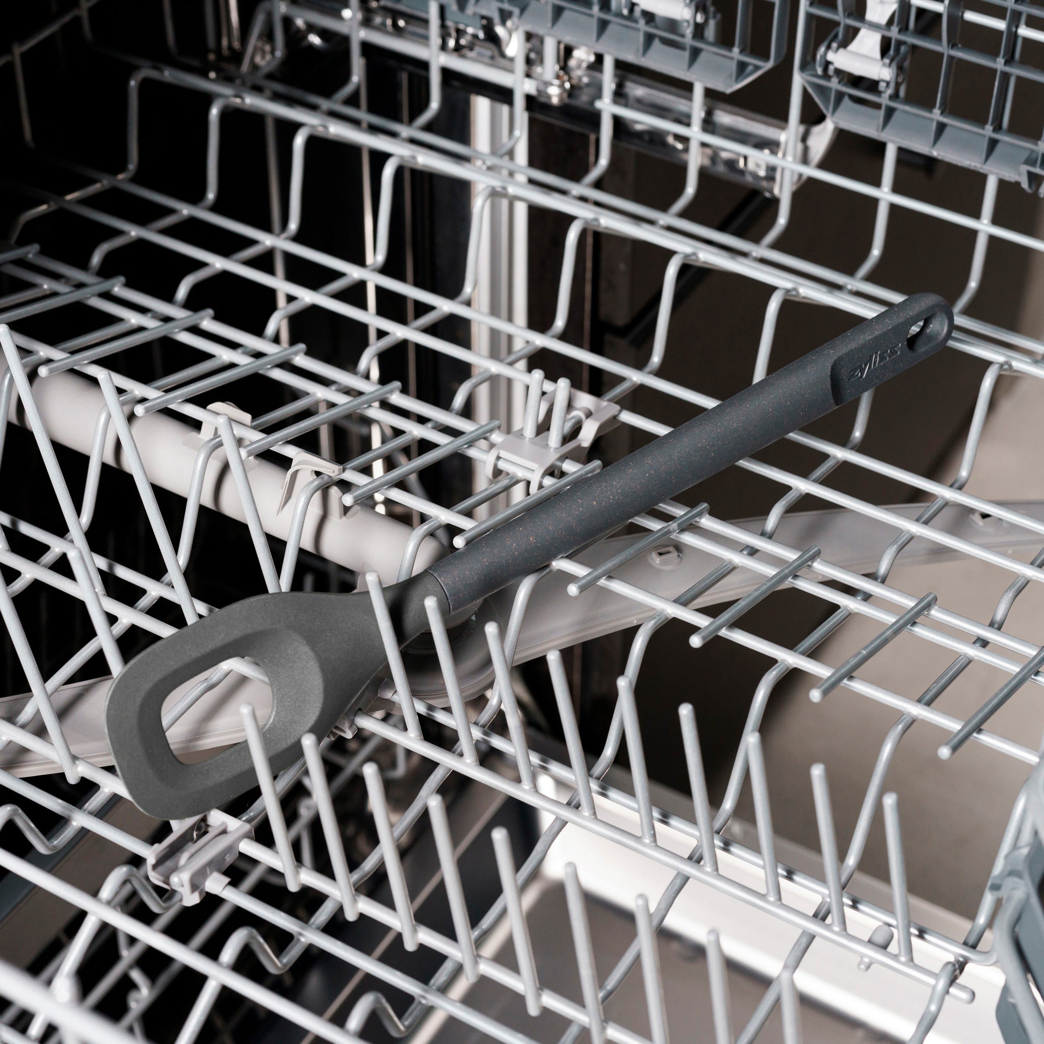 A black silicone spatula lies diagonally on the bottom rack of an open dishwasher, surrounded by the metal prongs of the rack.