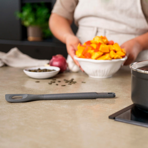 In a kitchen, a person in an apron holds a bowl of chopped orange and yellow vegetables. On the counter, a Zyliss Angled Mixing Spoon with an ergonomic handle rests beside non-stick cookware and a small bowl of seeds. 