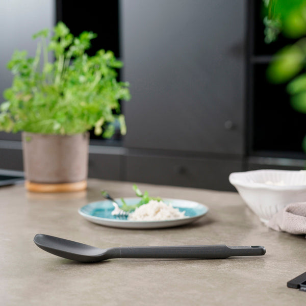 A Zyliss Spoon sits on a kitchen counter, with a green potted plant, a plate of rice and fork, and a white bowl in the background. #Medium