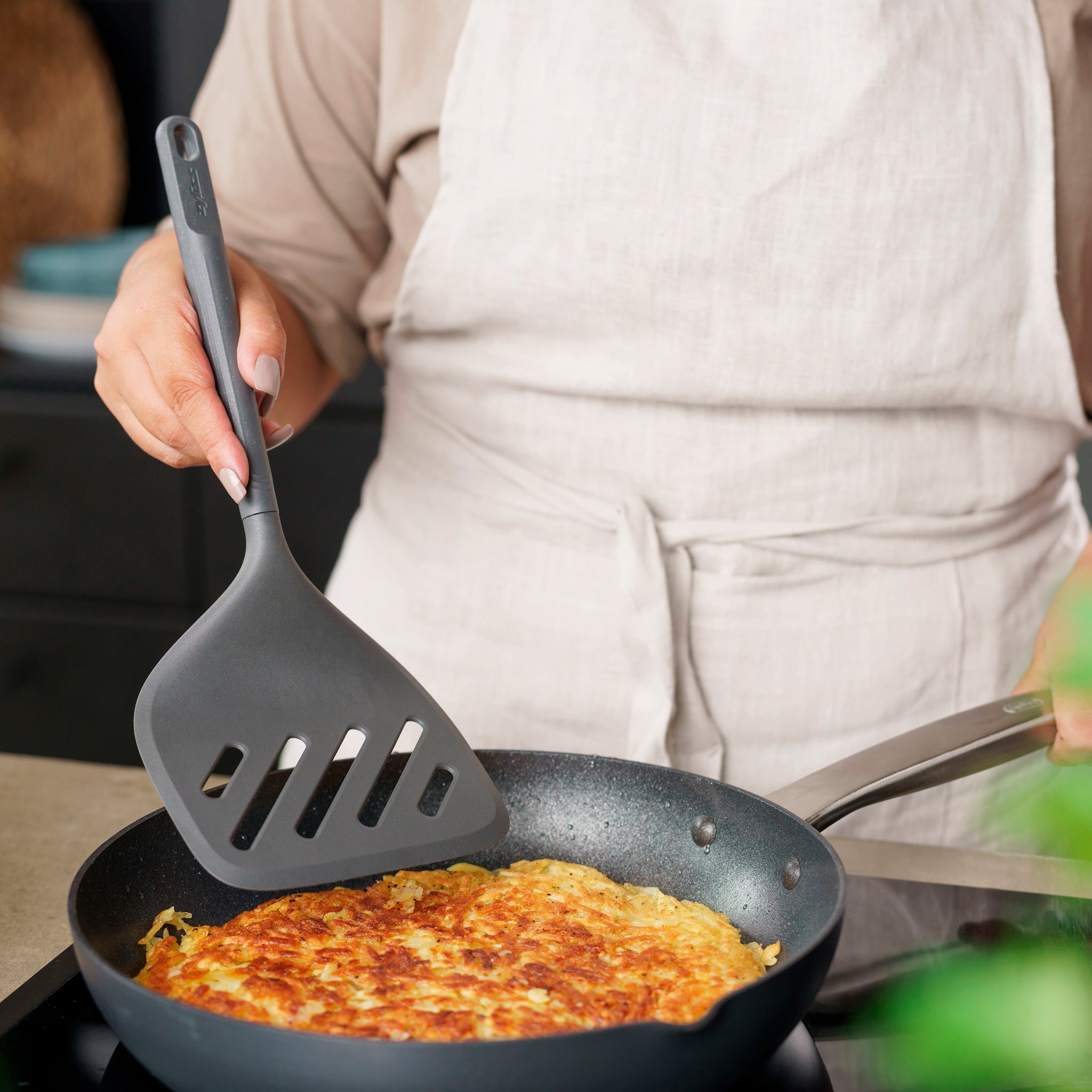 Wearing a light apron, a person uses the Zyliss Turner XL to lift or flip a golden-brown hash brown in a nonstick frying pan on the stove. 