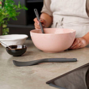 In a modern kitchen, someone in an apron uses a heat-resistant spatula to mix ingredients in a pink bowl while the Zyliss Easy Lift Turner rests on the counter among small bowls and a plant. 