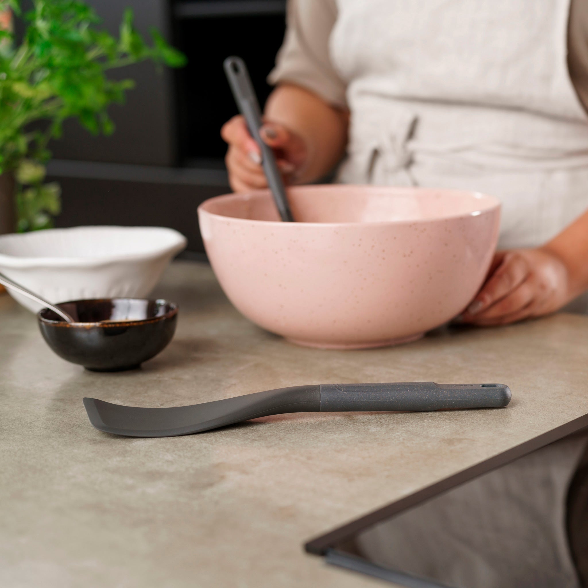 In a modern kitchen, someone in an apron uses a heat-resistant spatula to mix ingredients in a pink bowl while the Zyliss Easy Lift Turner rests on the counter among small bowls and a plant. 