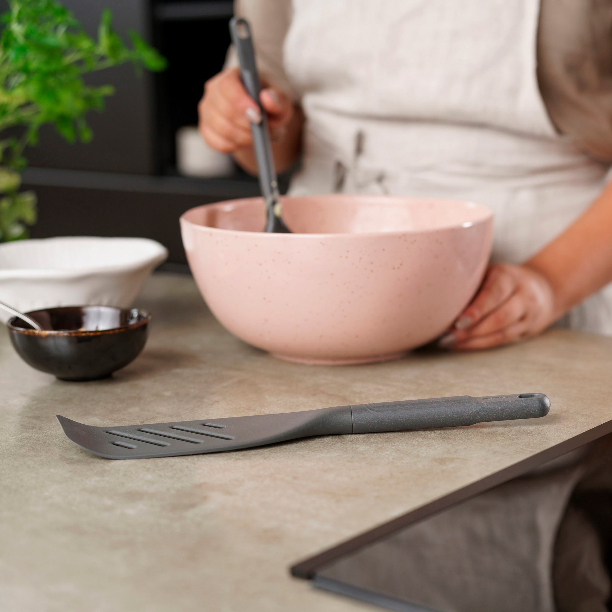 A person in an apron stands at the kitchen counter, mixing ingredients in a large pink bowl. A Zyliss Fish Turner rests nearby, along with a small white bowl and a plant visible in the background. 