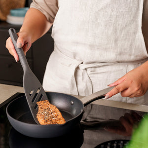 Wearing a beige apron, a person uses the Zyliss Fish Turner, which is heat-resistant and safe for nonstick cookware, to cook seasoned salmon in a nonstick frying pan on a stovetop. 