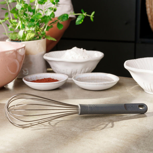 A Zyliss Balloon Whisk with a gray silicone handle sits on a kitchen counter, safe for non-stick cookware. It’s surrounded by bowls of flour and cocoa powder, with a plant and part of a person’s arm in the background. #Large