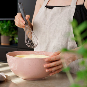 Wearing a light linen apron, someone uses the Zyliss Balloon Whisk to mix batter in a large pink bowl on the kitchen counter, surrounded by green plants. Its heat-resistant silicone is ideal for non-stick cookware and safe baking. #Large