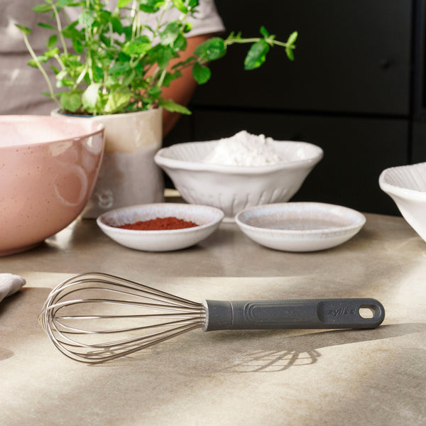 A Zyliss Balloon Whisk, made of heat-resistant silicone, sits on a kitchen counter amid a pink mixing bowl and small dishes—ideal for non-stick cookware. #Medium