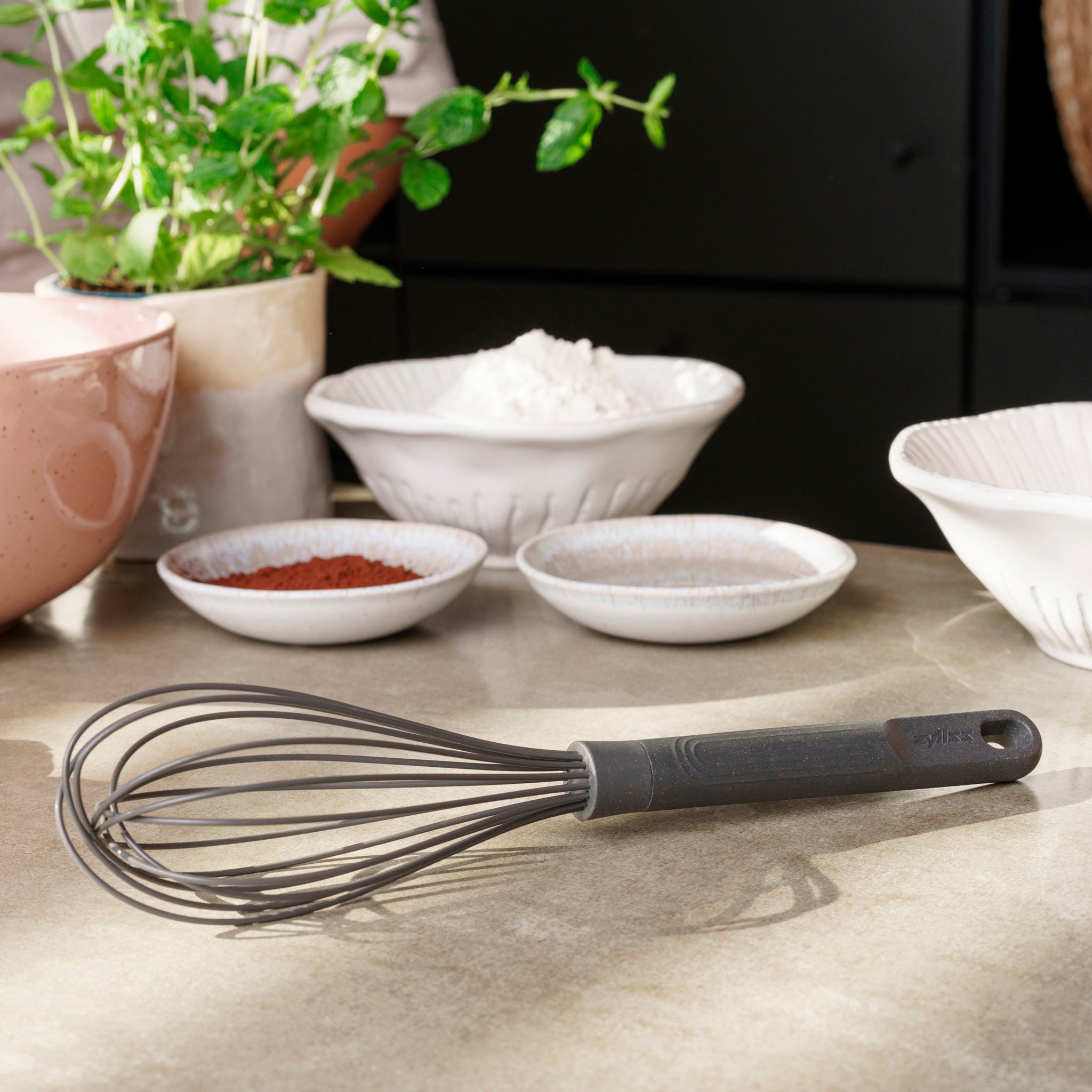 The Zyliss Balloon Whisk Silicone, safe for non-stick cookware, sits on a kitchen counter with bowls of flour, cocoa powder, and sugar nearby, next to a potted green plant. 