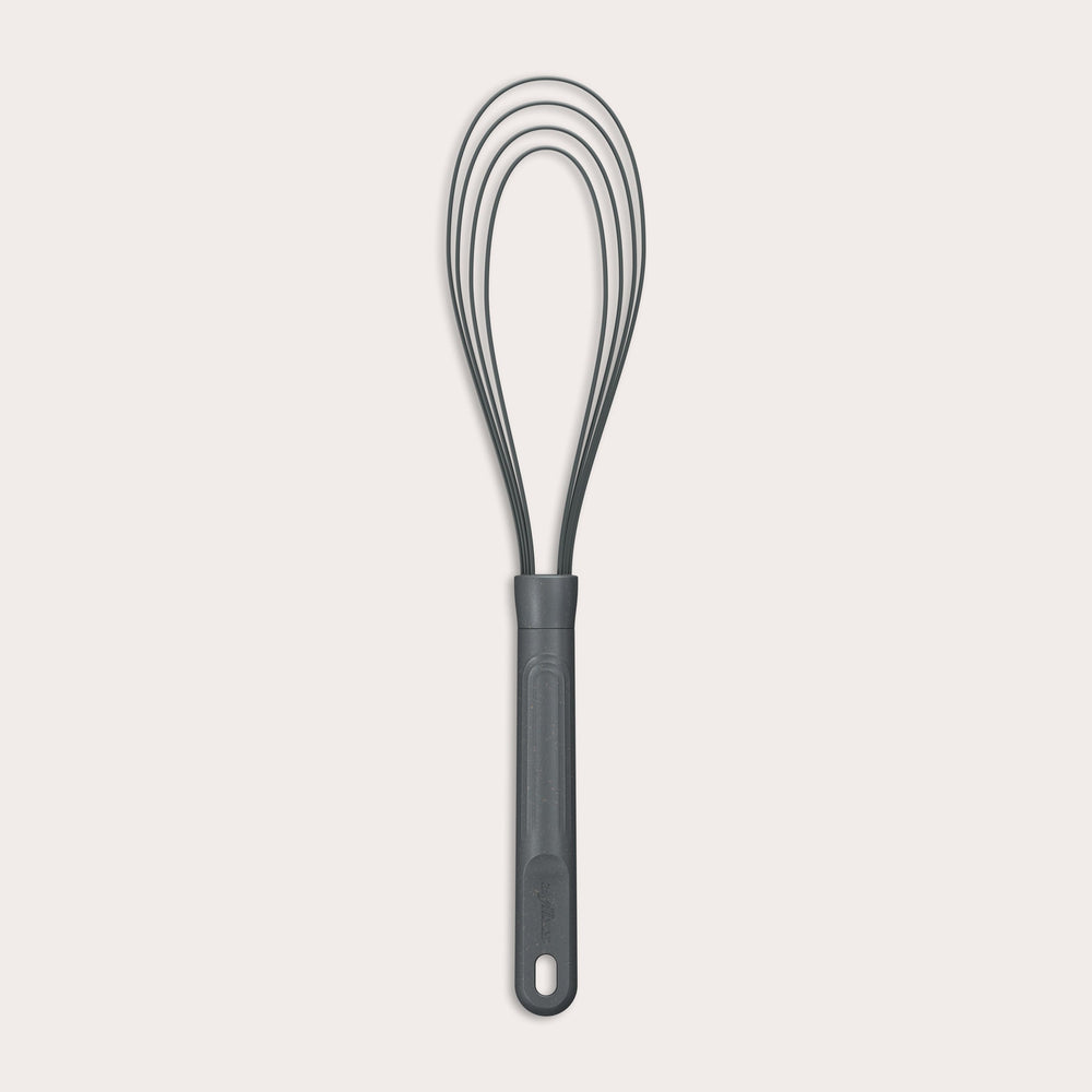 The Zyliss Flat Whisk Silicone features a black, heat-resistant silicone design with a long handle and looped wires, displayed on a plain light background. 