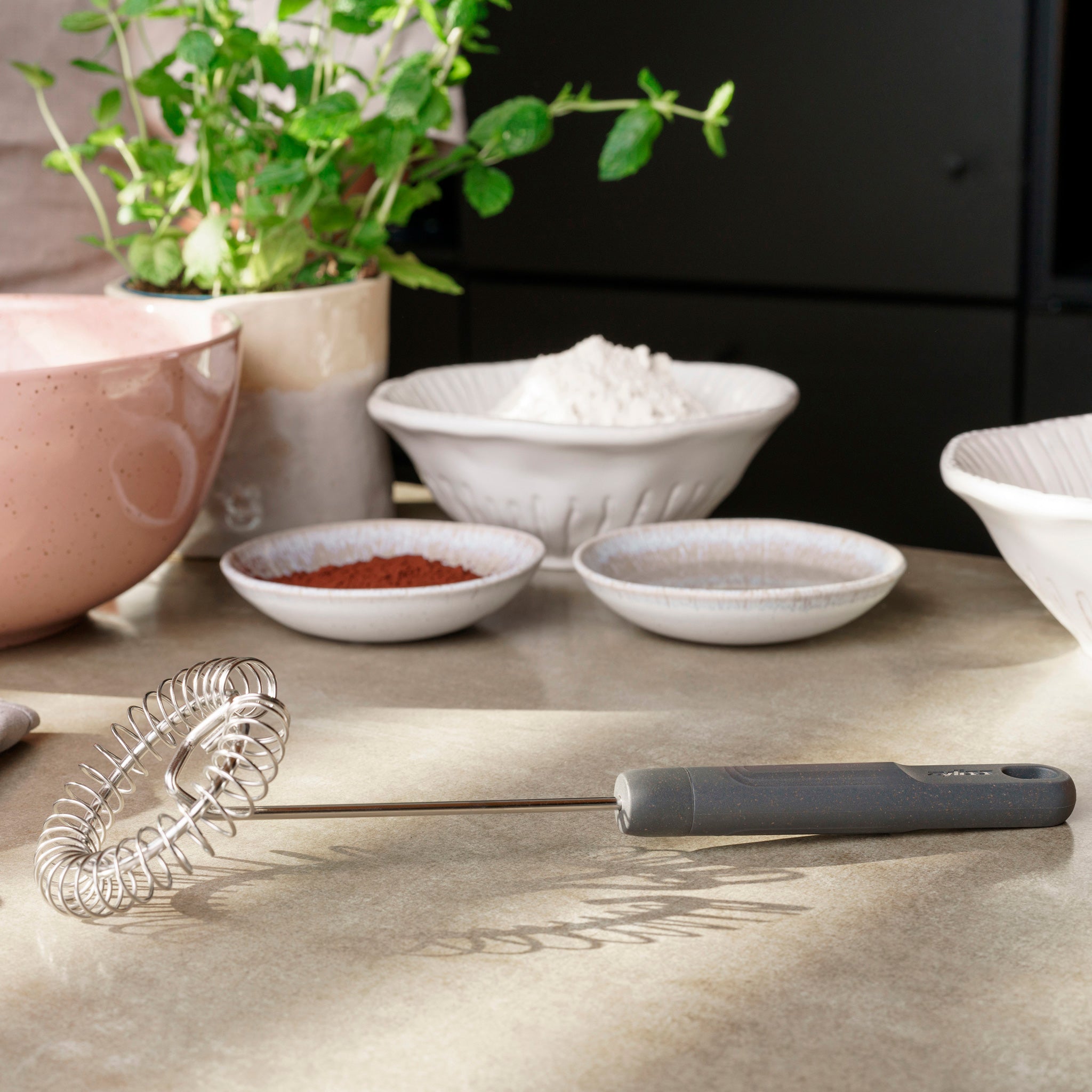 A Zyliss Gravy Whisk rests on a kitchen countertop with bowls of cocoa powder, flour, and sugar for sauce prep; a pink mixing bowl and a green potted herb are in the background. 