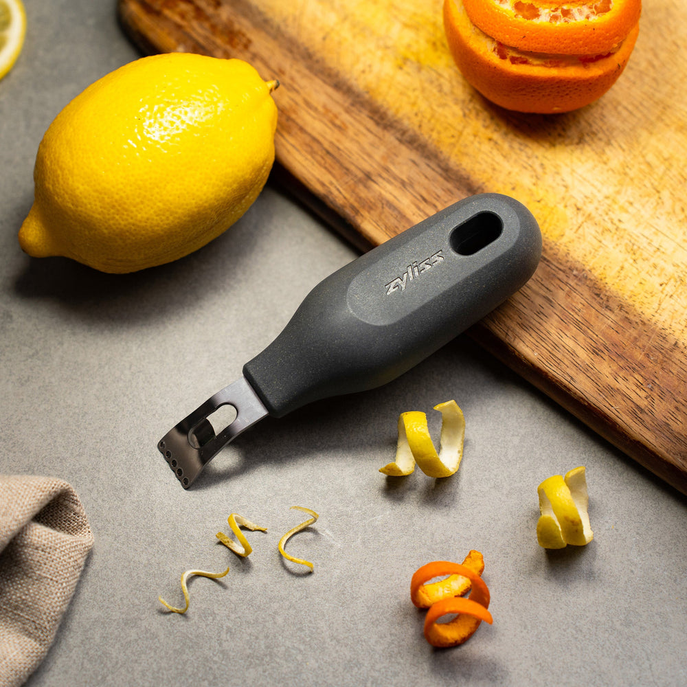 The Zyliss 2-In-1 Zester, made of stainless steel with a black ergonomic handle and a hole at the base, is an eco-friendly kitchen tool shown against a plain light background. 