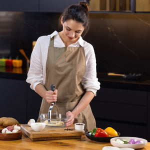 A woman in a beige apron uses the Zyliss Potato Masher with a stainless steel head to mash potatoes in a glass bowl on the kitchen counter, surrounded by colorful bell peppers, garlic, onions, and ingredient bowls as she smiles. 