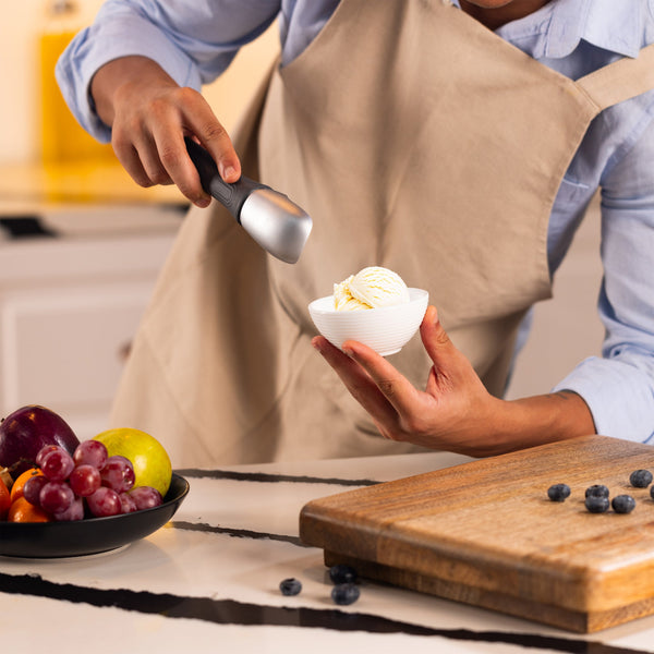#belugagrey Wearing a beige apron, a person uses the Zyliss Ice Cream Scoop with an eco-friendly handle to serve vanilla ice cream into a white bowl. Nearby are assorted fruit on a plate and blueberries on a wooden cutting board. 