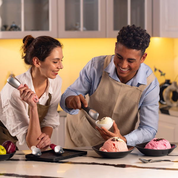 #naturalbeige A man and woman in aprons smile as they use the Zyliss Ice Cream Scoop, featuring a sustainable wheat straw, eco-friendly handle, to serve various flavors into bowls on the counter.