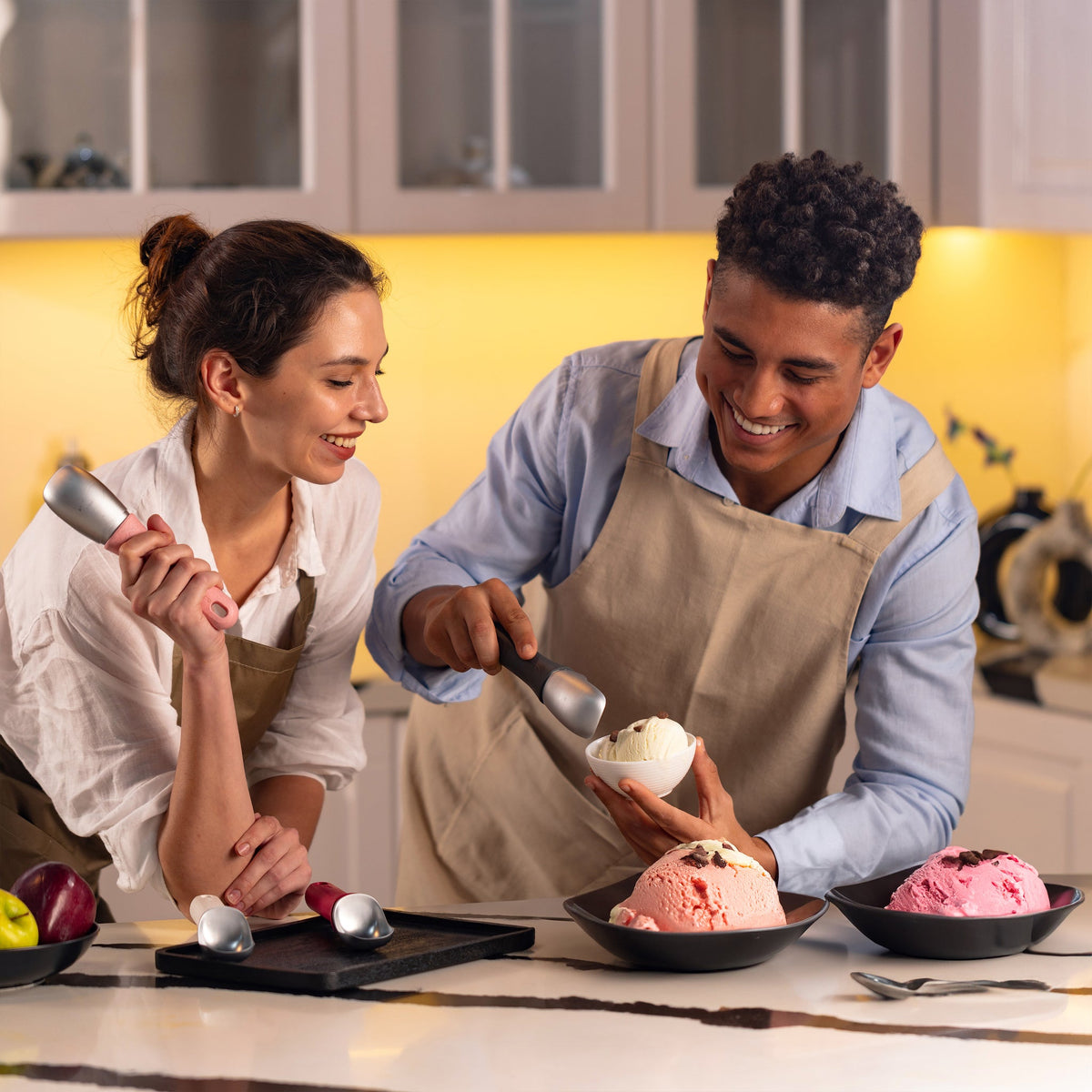#pink Two people in a kitchen smile as they use the Zyliss Ice Cream Scoop to serve white, pink, and light brown ice cream into bowls. Three large bowls of ice cream sit on the counter.