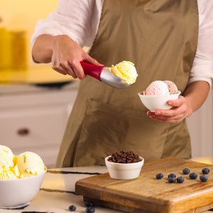 #berry Wearing a beige apron, a person uses the Zyliss Ice Cream Scoop to serve vanilla ice cream into a small bowl. Other bowls of ice cream, chocolate chips, a cutting board, and scattered blueberries are on the counter.