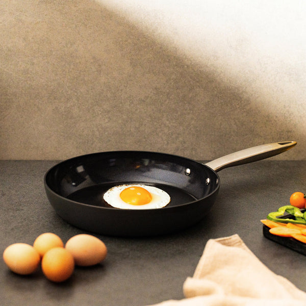 The Zyliss Ultimate Pro Ceramic Fry Pan with a non-stick coating holds a sunny-side-up egg on a dark countertop. Three brown eggs are nearby, along with cut vegetables on a tray and a beige cloth in the foreground. #24cm/9½"