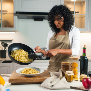 In a modern kitchen, a woman in an apron pours cooked pasta from the Zyliss Ultimate Pro Ceramic Fry Pan onto a plate, surrounded by jars of ingredients, a wine bottle, and cooking utensils. #28cm/11"
