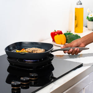 A hand holds the Zyliss Ultimate Pro Ceramic Square Grill Pan on a gas stove, cooking chicken and vegetables. On the counter are bell peppers, broccoli, a small plant, and oil in a clean, modern kitchen. 