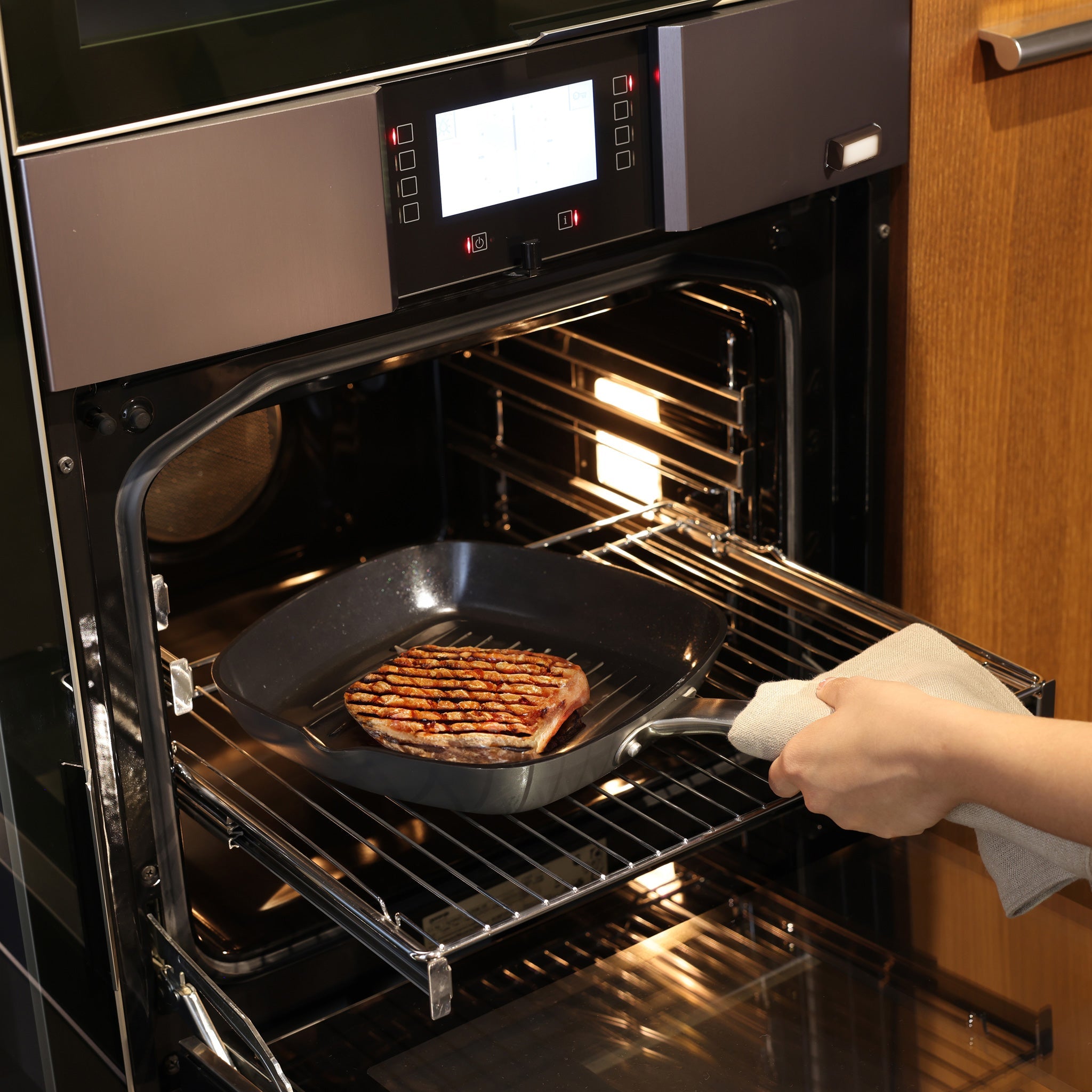 A person places a grill pan with a steak into an open oven, holding the pan handle with a cloth, while the oven’s interior light is on.