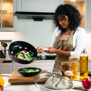 A woman in an apron cooks in a modern kitchen, pouring sautéed vegetables from a Zyliss Ultimate Pro Ceramic Saute Pan With Lid into a bowl. The counter holds fresh produce, jars, oil, and various kitchen items. 