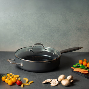 A Zyliss Ultimate Pro Ceramic Saute Pan With Lid rests on a gray counter, with uncooked pasta, cherry tomatoes, mushrooms, sliced carrots, and green bell peppers around it. 