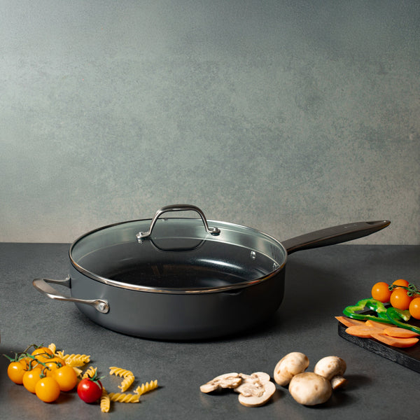 A Zyliss Ultimate Pro Ceramic Saute Pan With Lid rests on a gray counter, with uncooked pasta, cherry tomatoes, mushrooms, sliced carrots, and green bell peppers around it. 