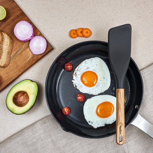 A frying pan with two sunny-side-up eggs and cherry tomatoes sits on a counter next to a Zyliss Oak Wood Easy Lift Turner, half an avocado, sliced red onion, carrot, whole wheat toast, and lime wedges on a wooden board.