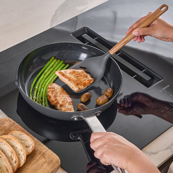 A person uses the Zyliss Oak Wood Slotted Turner to cook chicken breast, green beans, and small potatoes in a black frying pan on the stovetop, with slices of bread resting on an oak board for a classic kitchen moment.