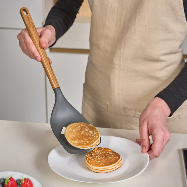 Wearing a beige apron, a person uses the Zyliss Oak Wood Slotted Turner to transfer small pancakes onto a white plate, with a bowl of berries visible in the foreground.