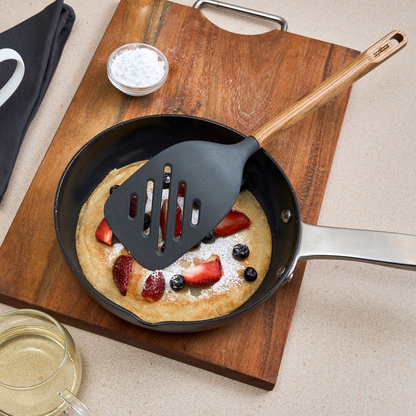 A pancake with strawberries, blueberries, and powdered sugar sits in a small skillet on an oak cutting board, with the Zyliss Oak Wood Slotted Turner resting on top and a glass bowl of syrup nearby.