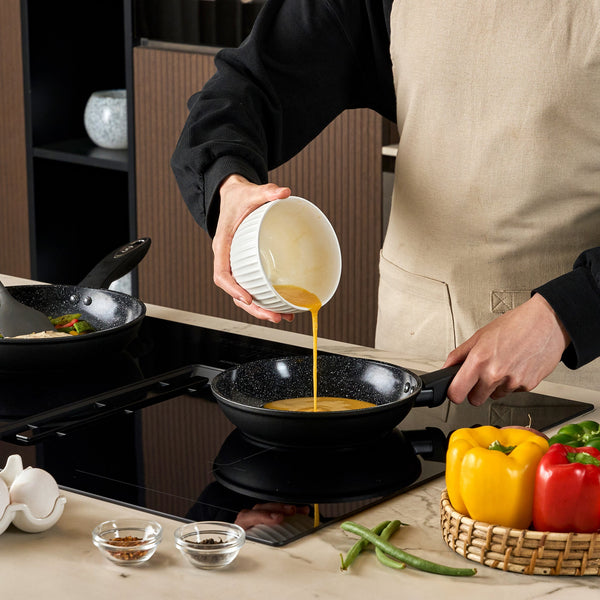 #20cm/8"
Wearing a beige apron, a person pours beaten eggs from a white bowl into the Zyliss Ultimate Ceramic Fry Pan on the stove. Nearby are bell peppers, green beans, eggshells, and bowls of spices.
