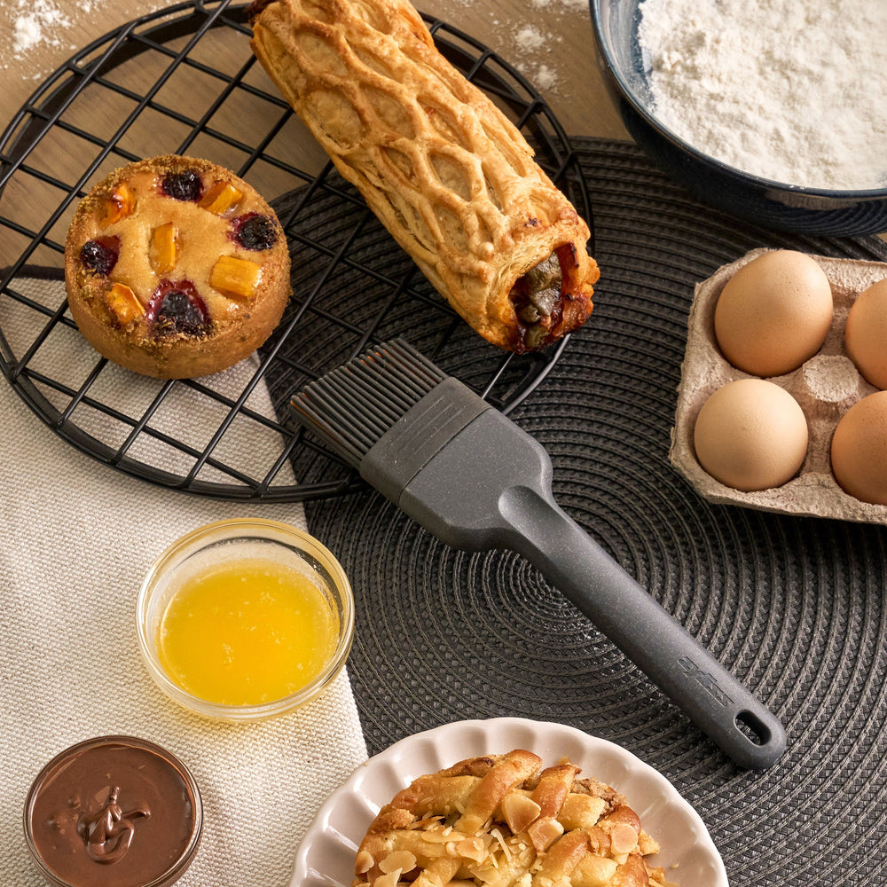 The Zyliss Silicone Pastry Brush is gray, heat-resistant, and dishwasher safe. It features a long handle with a hanging hole and flexible bristles ideal for spreading sauces or oils. Shown against a plain, light-colored background.