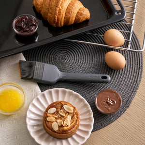 A heat-resistant Zyliss Silicone Pastry Brush beside a croissant on a baking tray, two eggs, an almond-topped pastry on a plate, and small bowls of jam, melted butter, and chocolate spread on a placemat.