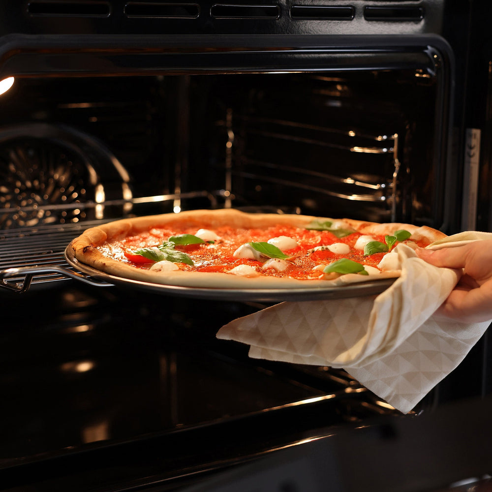 A Margherita pizza with tomato, mozzarella, and basil sits on a Zyliss Non-Stick Pizza Tray made of carbon steel, ensuring crispy bases. Basil, chili peppers, and a pizza cutter lie nearby on the wooden surface.