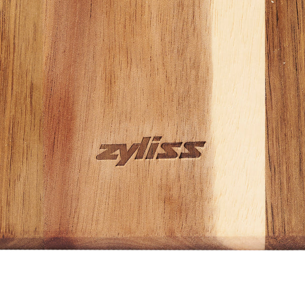 A close-up of the Signature 3 Piece Cheese Knife & Acacia Serving Board Set by Zyliss, featuring zyliss engraved in the center and showcasing striking light and dark brown acacia wood tones.