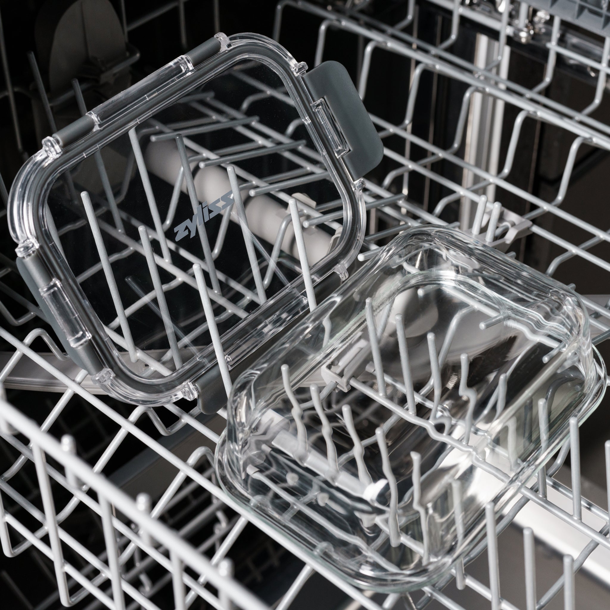 A clear, rectangular plastic food container with its lid open is placed on the top rack of a dishwasher. The container and lid are transparent and resting among the dishwasher’s white prongs.