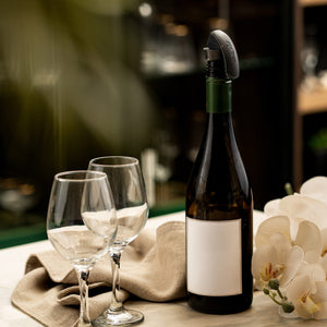 A wine bottle with a blank label and a Zyliss Bottle Stopper 2 Piece Set sits on a table near two empty glasses, a beige cloth, and white orchids. The softly blurred background enhances the elegant storage solution.