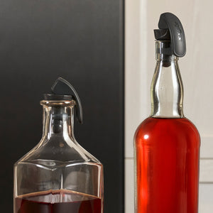 The Zyliss Bottle Stopper 2 Piece Set includes a stopper for a square glass decanter and another for a tall round bottle, both in black plastic—ideal for sleek, secure drink storage at home.