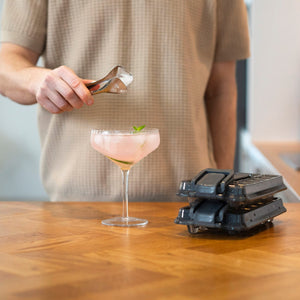 A person in a beige shirt uses metal tongs to place an ice cube from the Zyliss Press & Pull Ice Cube Tray into a pink cocktail in a coupe glass on a wooden countertop, with more trays stacked nearby.