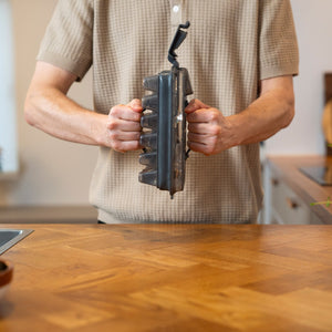A person in a beige short-sleeved shirt holds a waffle maker over a wooden counter, with the Zyliss Press & Pull Ice Cube Tray stacked nearby. The kitchen background is softly blurred.