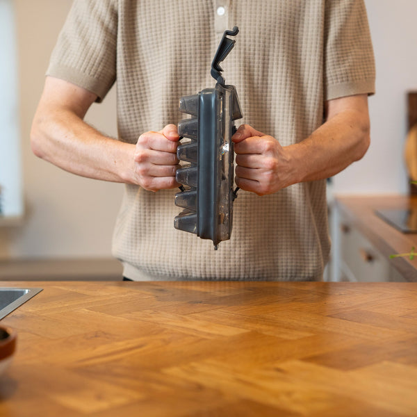 A person in a beige short-sleeved shirt holds a waffle maker over a wooden counter, with the Zyliss Press & Pull Ice Cube Tray stacked nearby. The kitchen background is softly blurred.