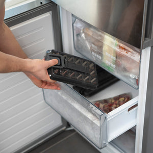 A person puts a black ice pack in the lower freezer drawer next to frozen foods and a Zyliss Press & Pull Ice Cube Tray, alongside a bag of frozen fruit or vegetables.