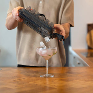 Someone pours ice cubes from the Zyliss Press & Pull Ice Cube Tray into a cocktail glass with a pink drink and lime slices, on a wooden kitchen counter.