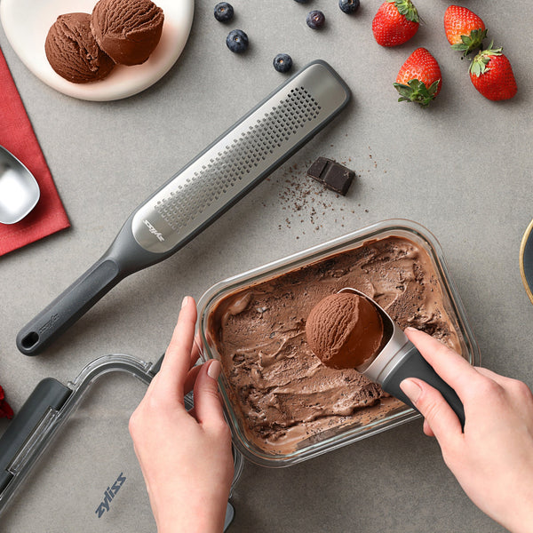 Someone scoops chocolate ice cream from a Zyliss Glass Container. Close by are strawberries, blueberries, a piece of chocolate, a metal grater, and a napkin on a gray surface—great for desserts or prepping meals. #1.52L/1.61qt