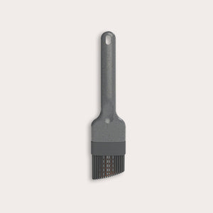The Zyliss Silicone Pastry Brush is gray, heat-resistant, and dishwasher safe. It features a long handle with a hanging hole and flexible bristles ideal for spreading sauces or oils. Shown against a plain, light-colored background.