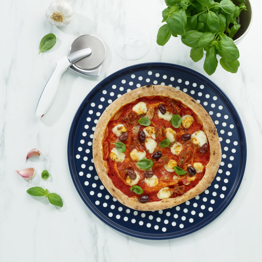 The Zyliss Durable Non-Stick Pizza Tray is a round, dark blue metal tray with raised edges and a perforated base featuring evenly spaced holes for optimal baking, shown against a plain light background.