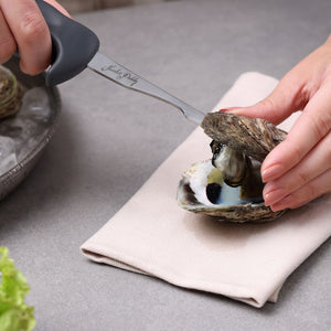 Using a Zyliss Oyster Knife Shucker Paddy, a person shucks oysters on a beige napkin, with a bowl of oysters on ice and lettuce nearby, all placed on a gray countertop.